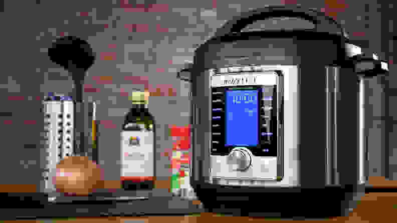 An Instant Pot shown besides some cooking utensils, an onion, an some sauces.