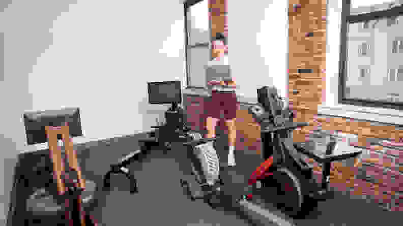 woman standing in a room with many different rowing machines and a laptop.