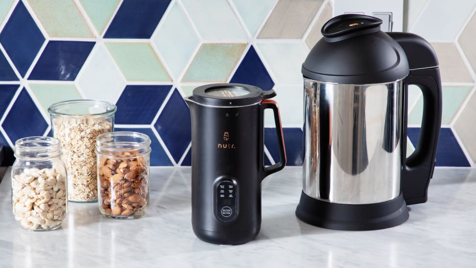 The Nutr plant-based milk maker next to the Almond Cow plant-based milk maker on granite countertop in front of tiled wall and 3 clear jars with snacks inside.