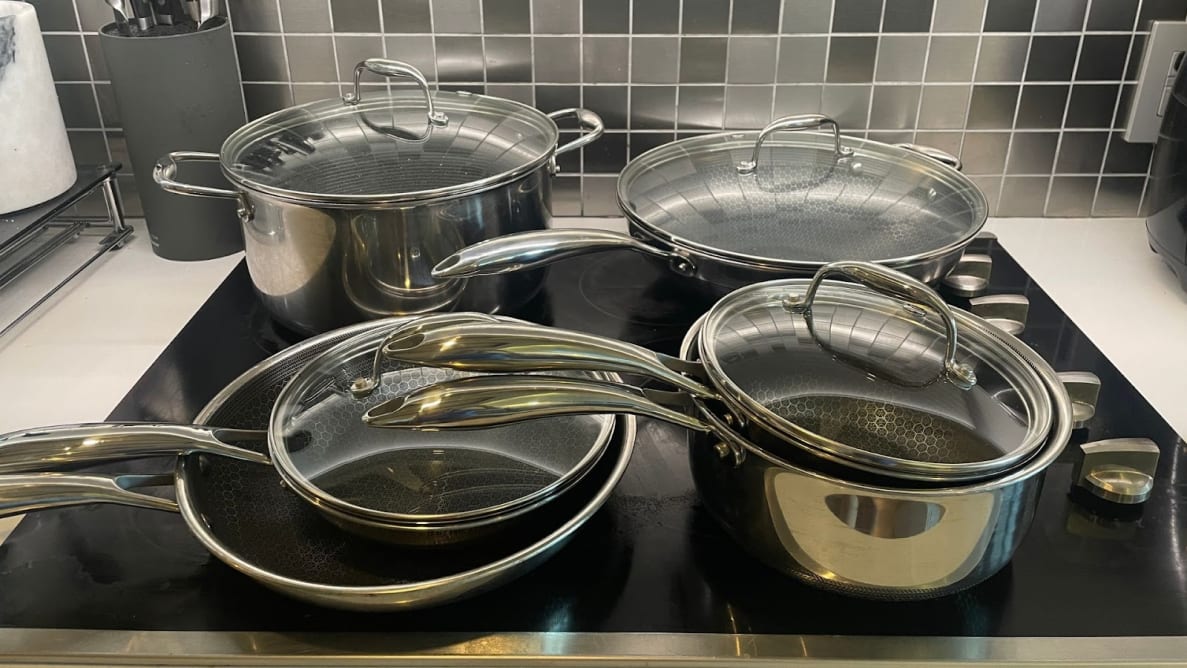 Stainless steel, nonstick Hexclad Hybrid cookware set sitting on top of stovetop with lids on top in front of tiled wall in kitchen.