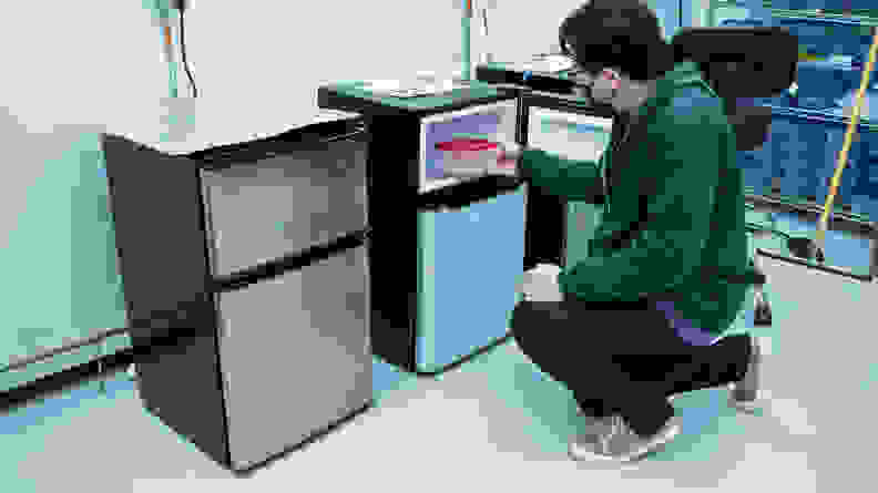 A man crouches by a mini fridge in the Reviewed labs, putting a container into the freezer portion of a mini fridge.