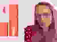 On the left: A coral-colored doe-foot applicator and lipstick tube. On the right: A person with pink hair wearing a red top and glasses purses their lips, which has a pink color on it.