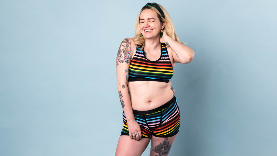 A person smiling in TomboyX bra and TomboyX 4.5-inch trunks against a blue backdrop.