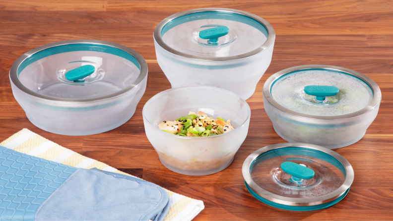 Four pieces of Anyday cookware laid out on a wooden surface next to two towels, one is open revealing leftover noodles.