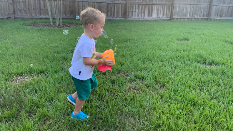 A toddler carries a bubble machine through the yard.