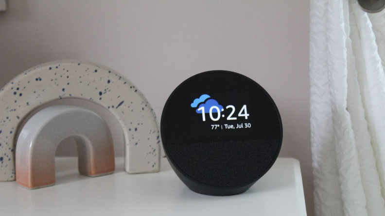 The Amazon Echo Spot on a nightstand, displaying the time and weather next to a small marble statue.