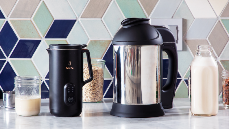 The Nutr plant-based milk maker next to the Almond Cow plant-based milk maker on granite countertop in front of tiled wall and 3 clear jars with snacks inside and one glass jar of milk.