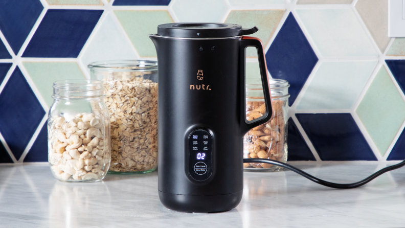 Black Nutr plant-based milk maker sitting on granite countertop in front of three glass jars with snacks inside.