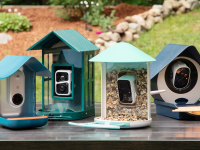 Four bird feeders in blue and green shades sit in a group on a brown table with a garden and green grass in the background