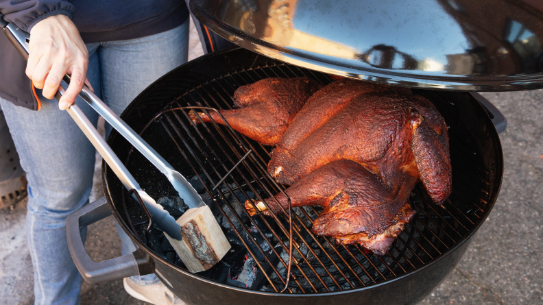 A person adds a block of wood to the charcoal grill while a turkey is smoking.