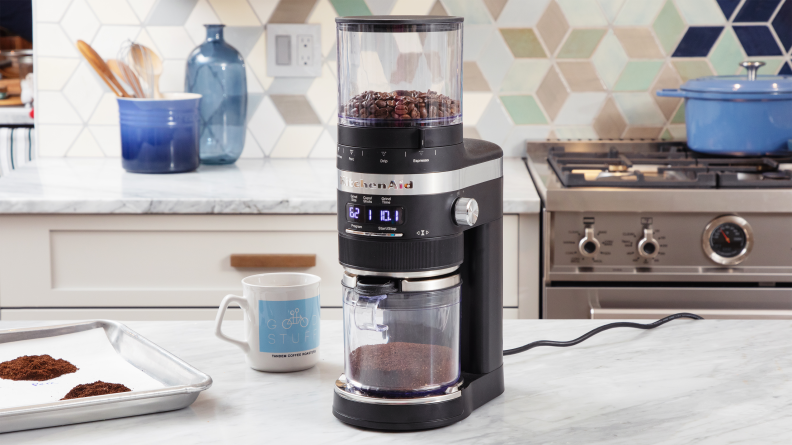 KitchenAid grinder on a countertop surrounded by a coffee cup and coffee grounds