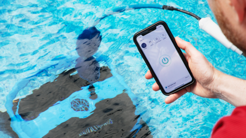 A person standing over a swimming pool with the Dolphin Nautilus CC Plus Wi-Fi in the water. The person is holding a smart phone with the Dolphin app showing.