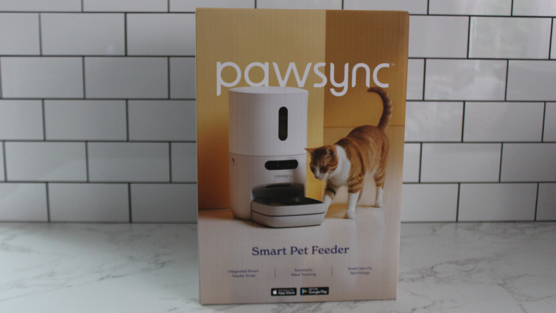 The box for the PawSync Smart Pet Feeder on a marble countertop in front of a tile wall.