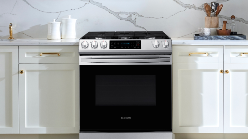 A stainless-steel Samsung slide-in gas range is surrounded by white cabinets with gold accents.