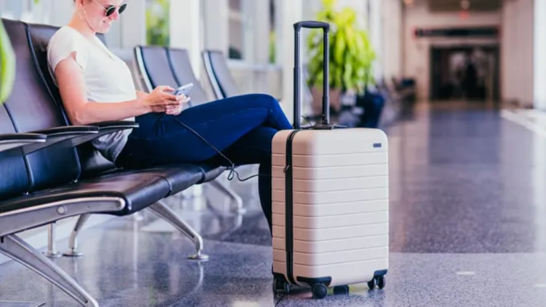 A woman sitting in an airport, looking at their phone, next to an Away Luggage bag.