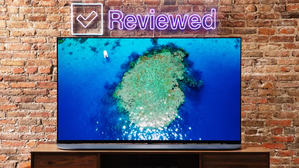 An OLED TV with an island on screen sits on a wooden desk before a brick wall with the neon sign that says "reviewed."